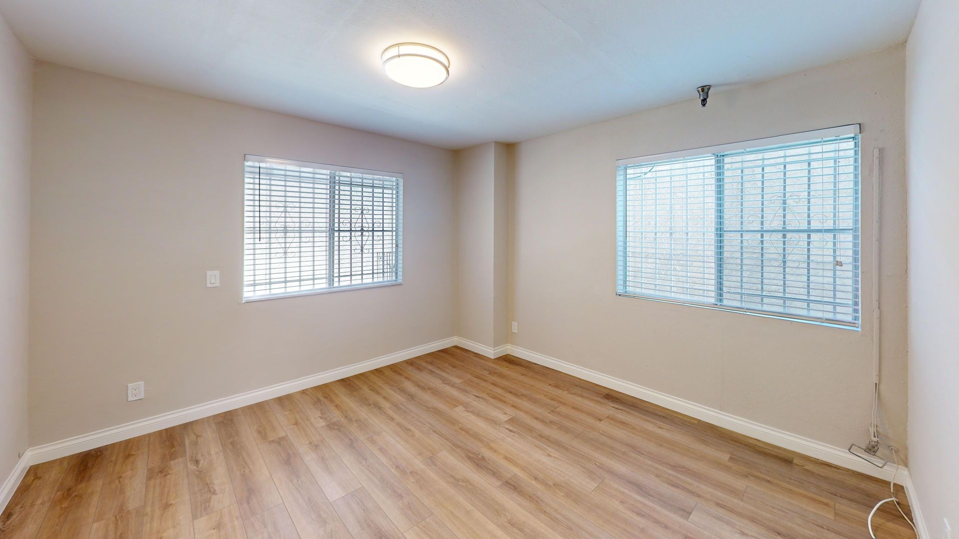Empty room with two windows, light-colored walls and wood flooring, ceiling light.