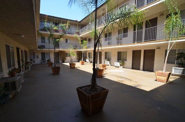 Apartment complex courtyard with potted trees, two-story building, doors and balconies, sunny day.