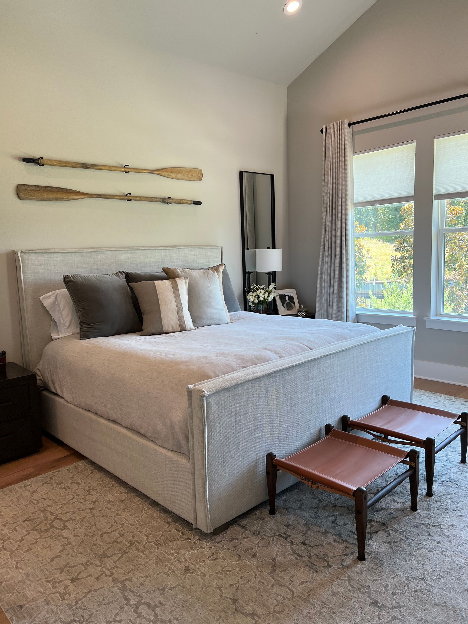 A bedroom with a large bed , two ottomans , a mirror and two windows.