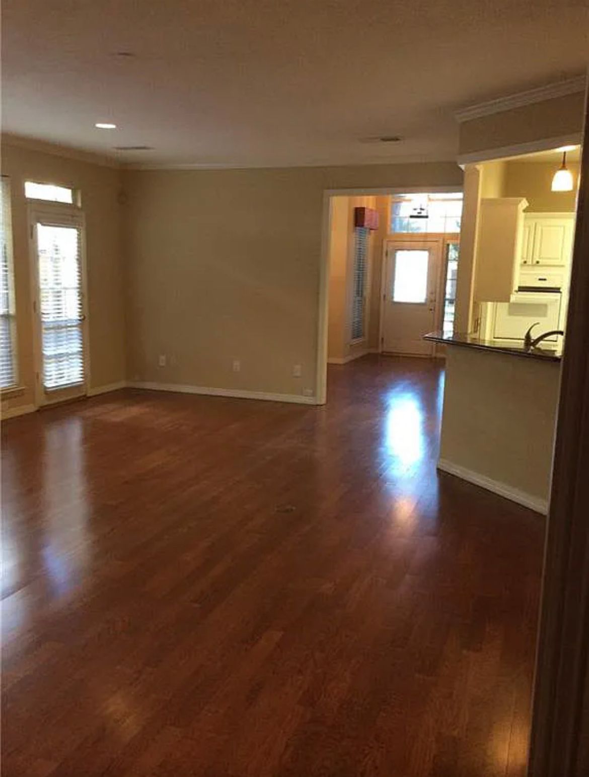An empty living room with hardwood floors and a kitchen in the background.
