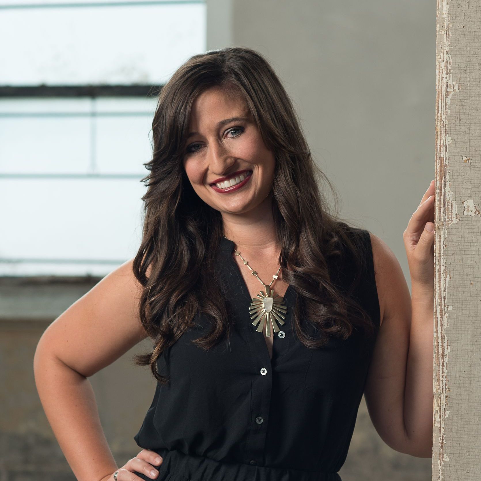A woman in a black dress is smiling and leaning against a wall