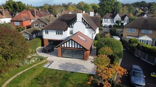 Suburban house with white facade, black trim, and attached garage. Brown roof, surrounded by trees.