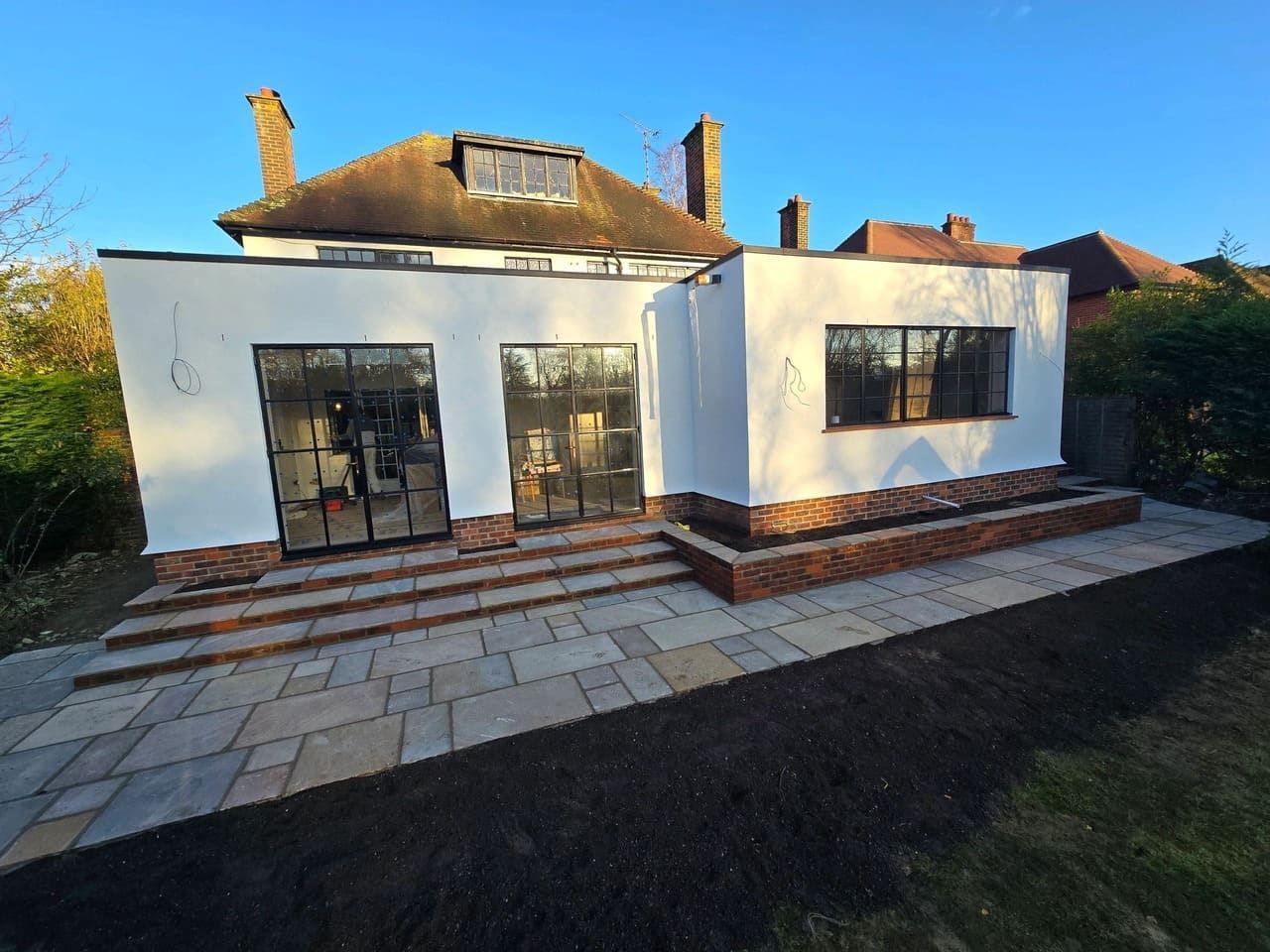 White and brick extension with steel doors, window, and stone patio. Sunlight, blue sky.
