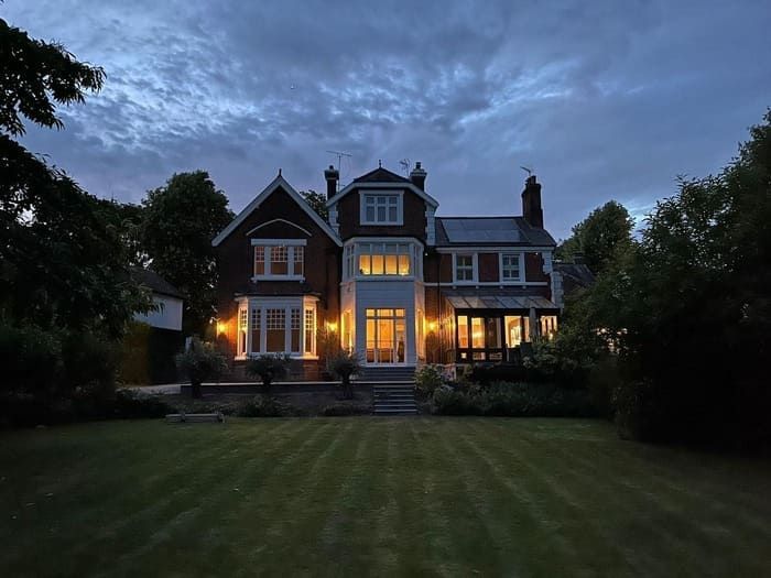 A large house at dusk with lit windows, set on a lawn with trees under a cloudy sky.