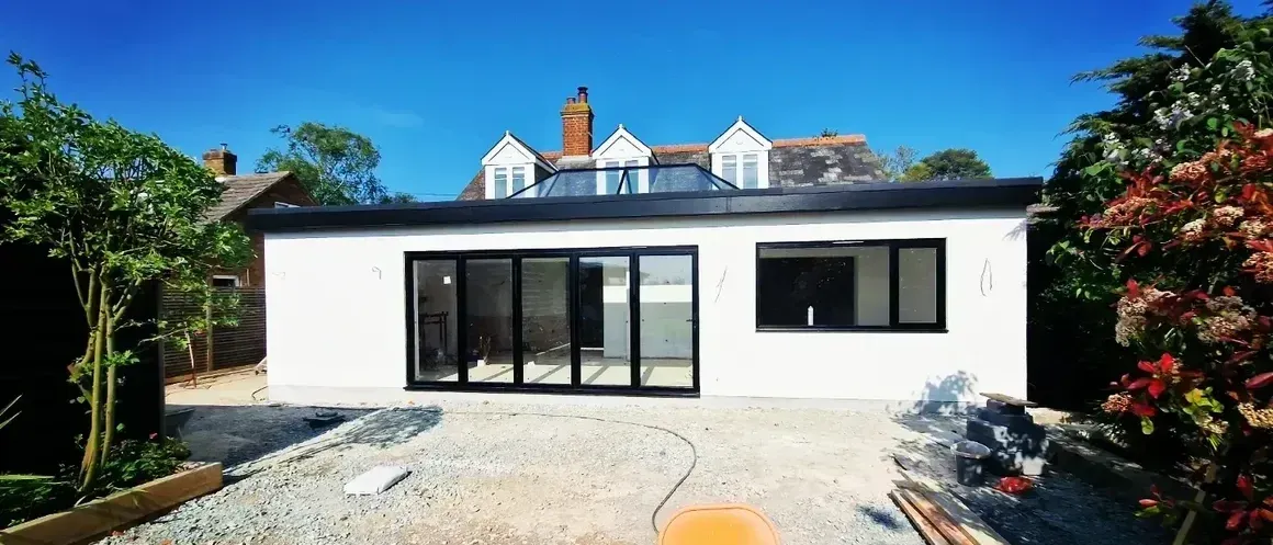 White building with black windows and doors, blue sky, gravel yard, and trees.