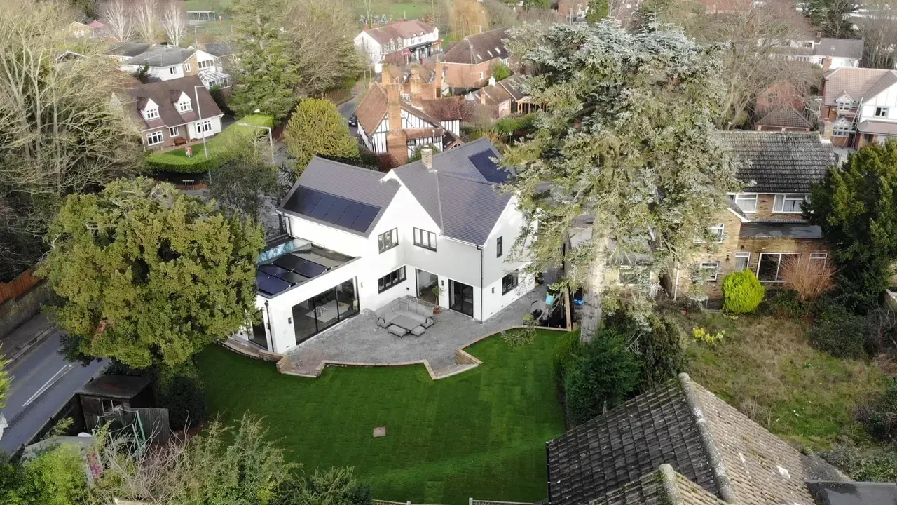 Aerial view of modern white house with black trim, patio, and green lawn. Surrounded by trees and other houses.
