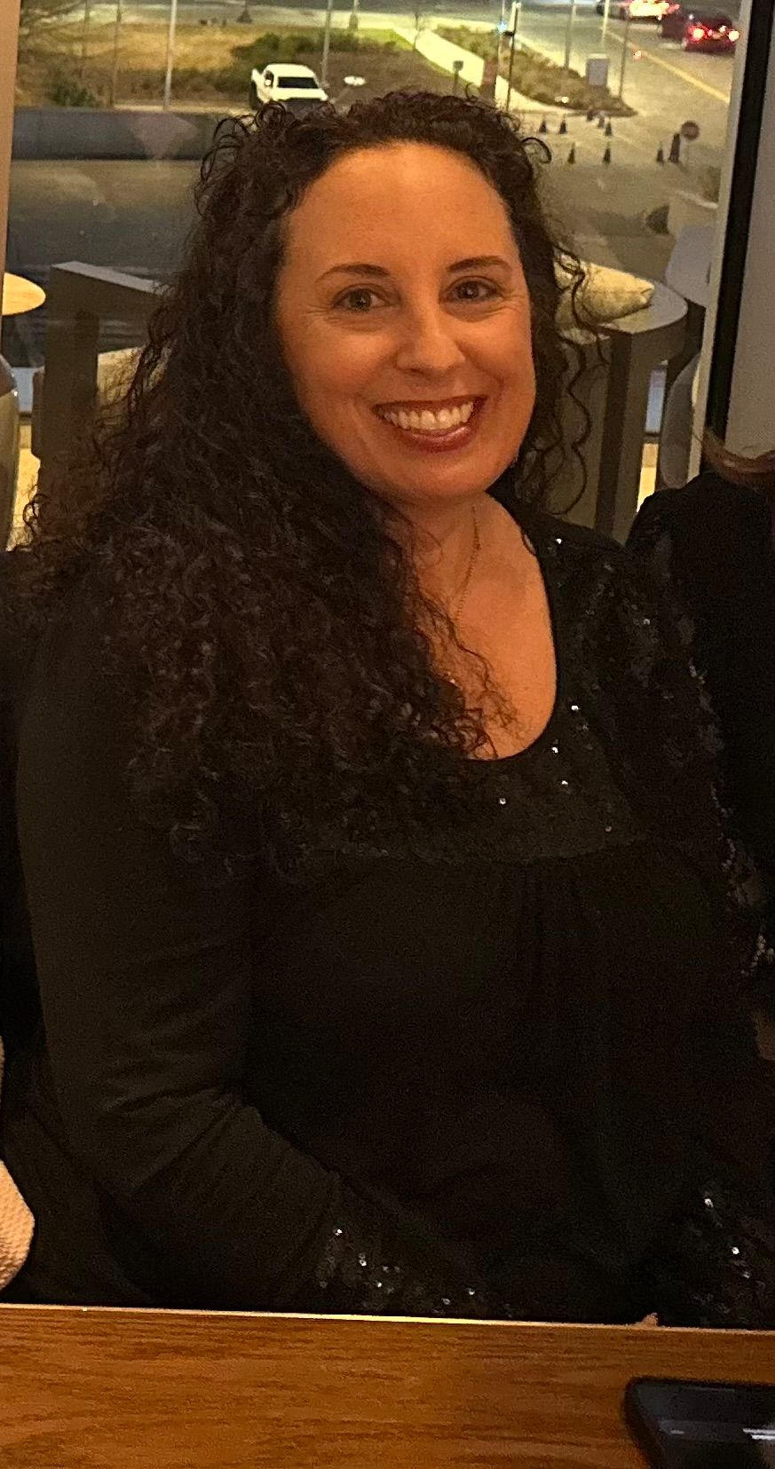 A smiling person with long dark curly hair, wearing a black top, seated indoors near a window.