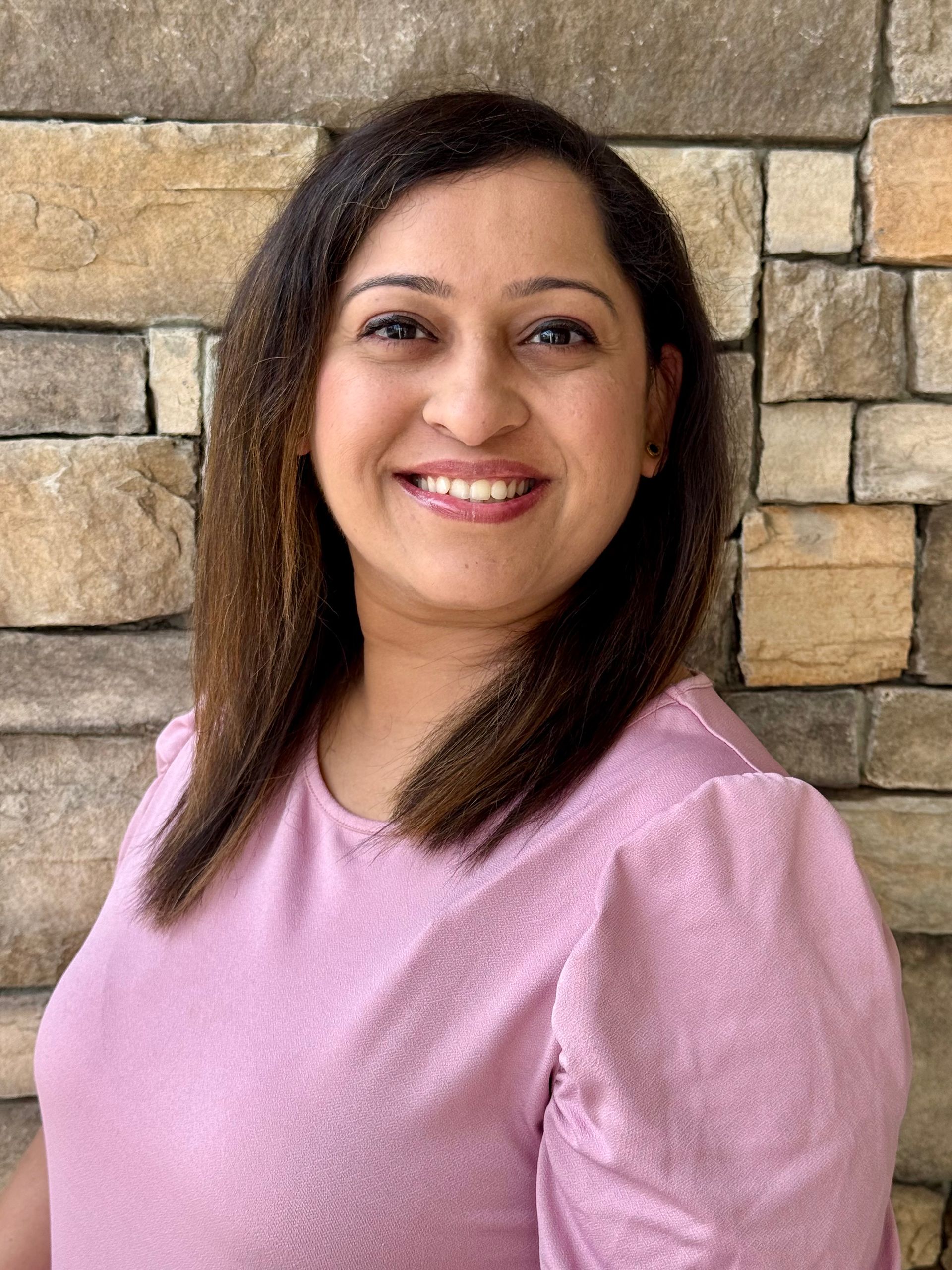 A smiling person with dark hair wearing a light pink shirt, positioned in front of a textured stone wall.
