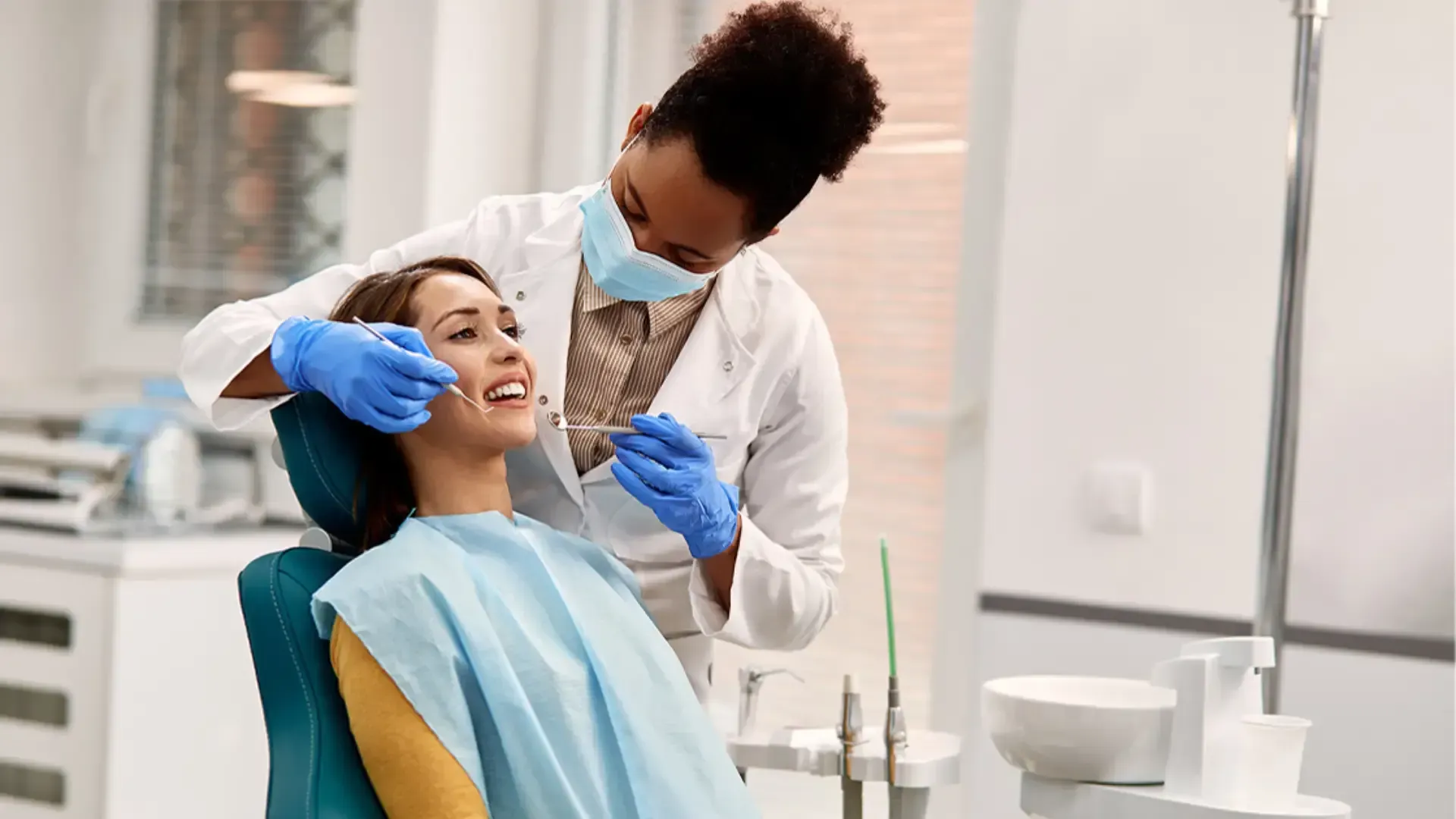 A masked dentist wearing blue gloves examines a smiling patient sitting in a dental chair.