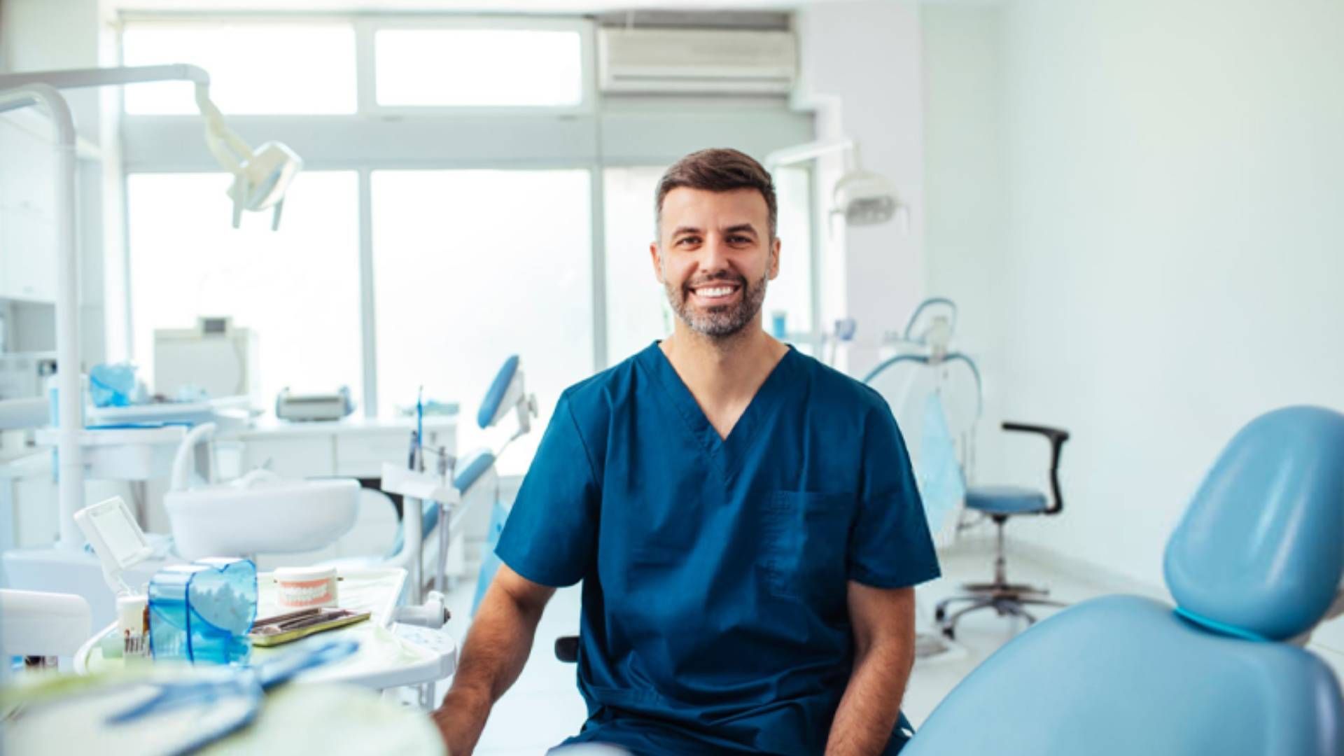 A smiling dental professional in navy blue scrubs sits in a brightly lit dental office.