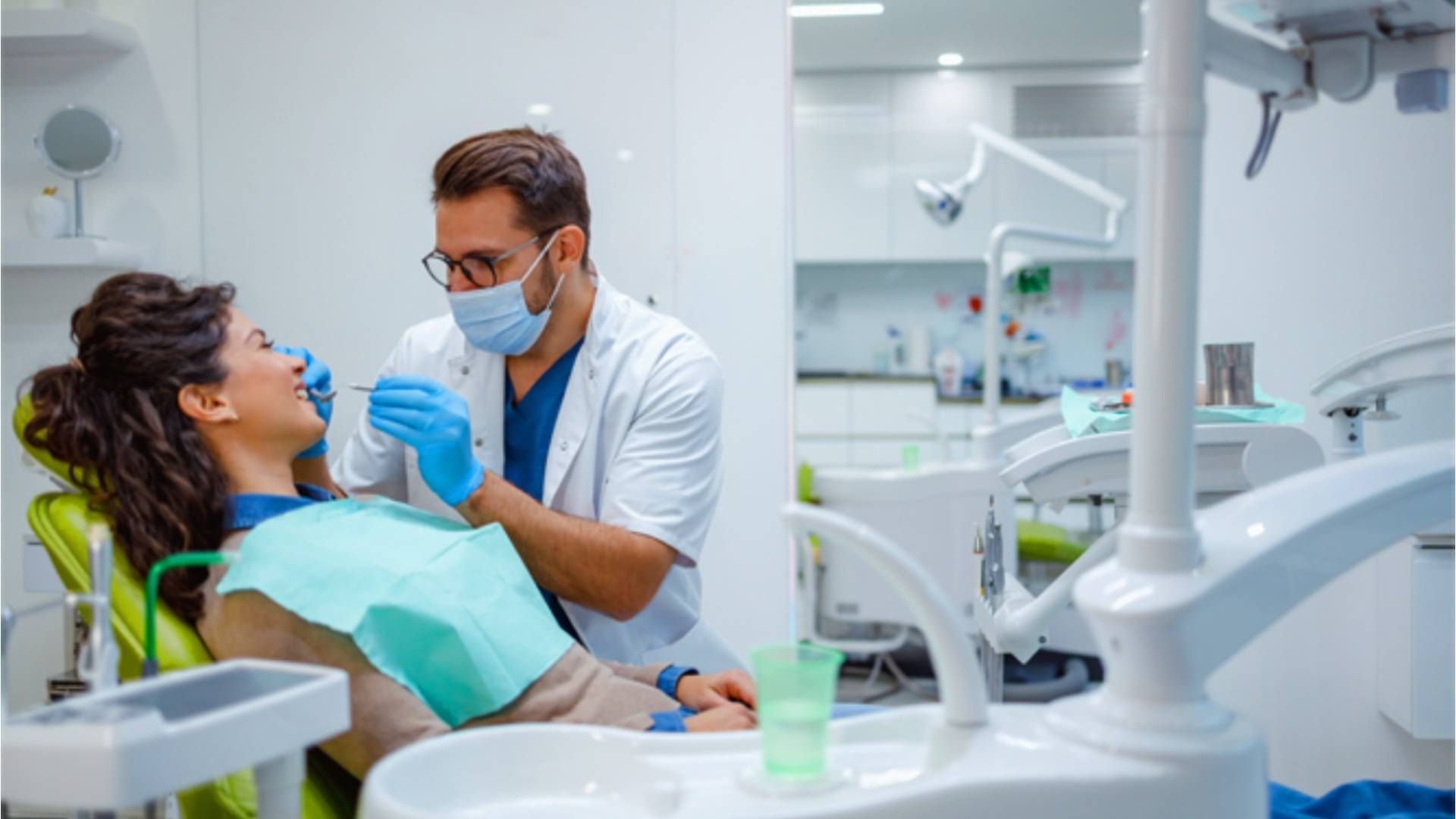 A dentist wearing a mask and gloves examines a patient sitting in a chair in a bright, modern clinic.