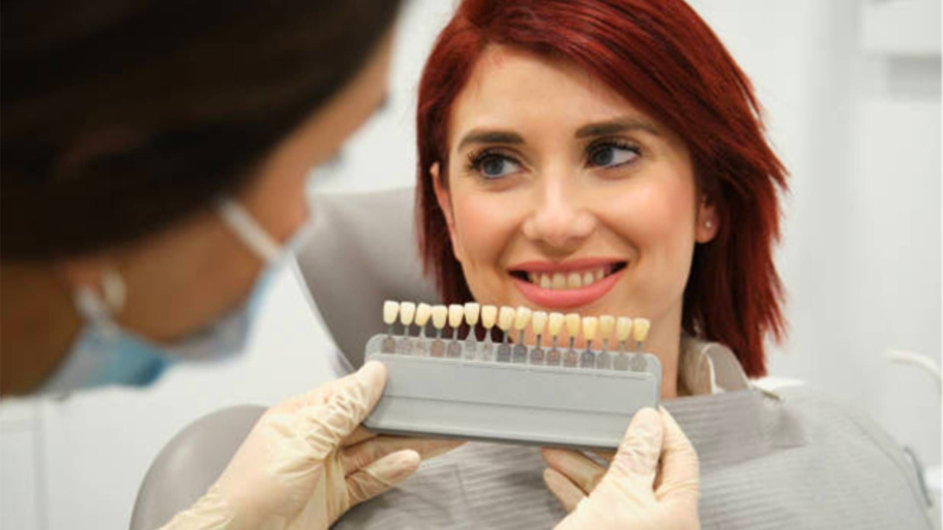 A dentist holding a tooth shade guide in front of a smiling patient in a dental office.