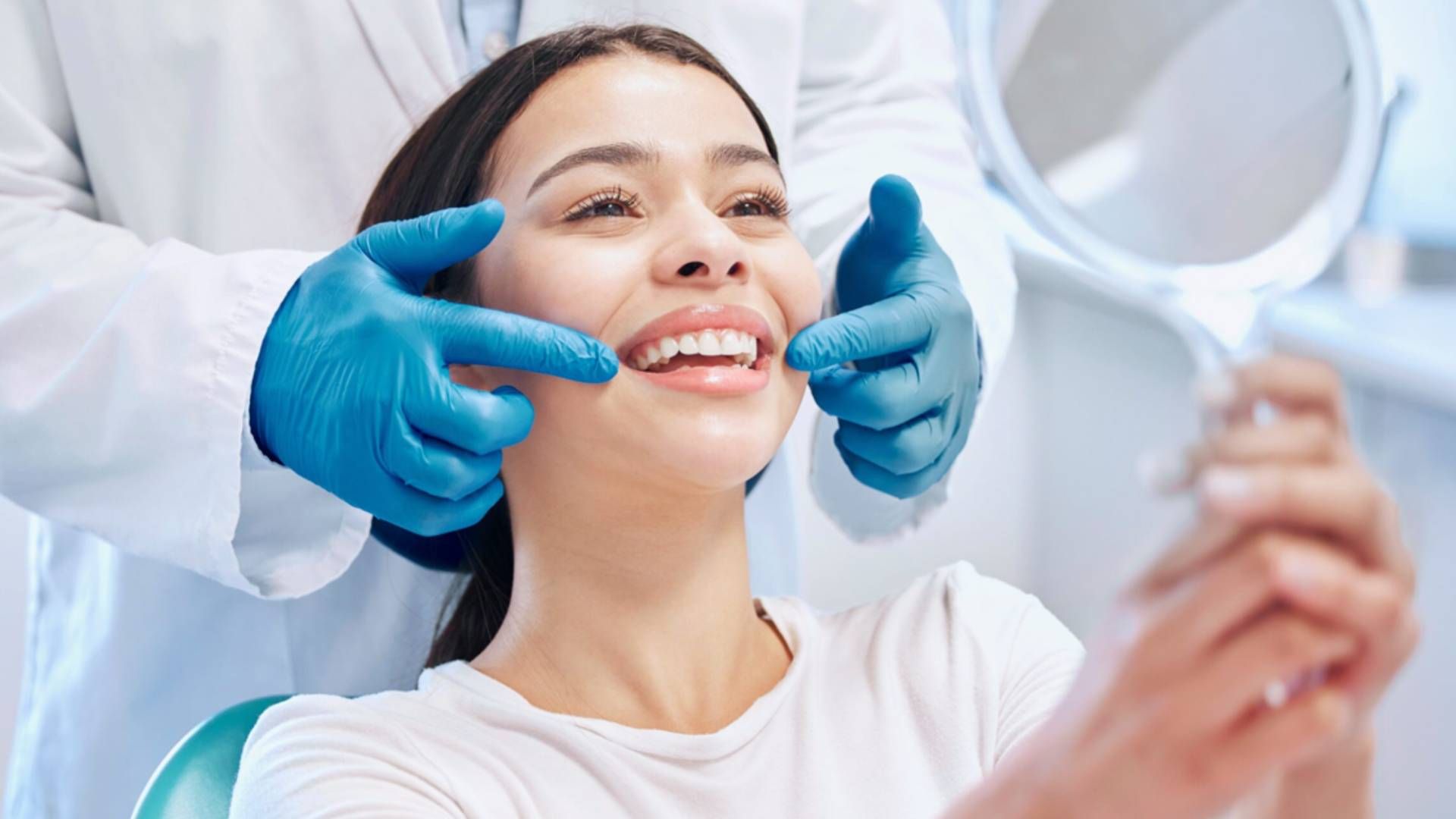 A dentist in blue gloves examines a smiling patient holding a mirror in a dental office.