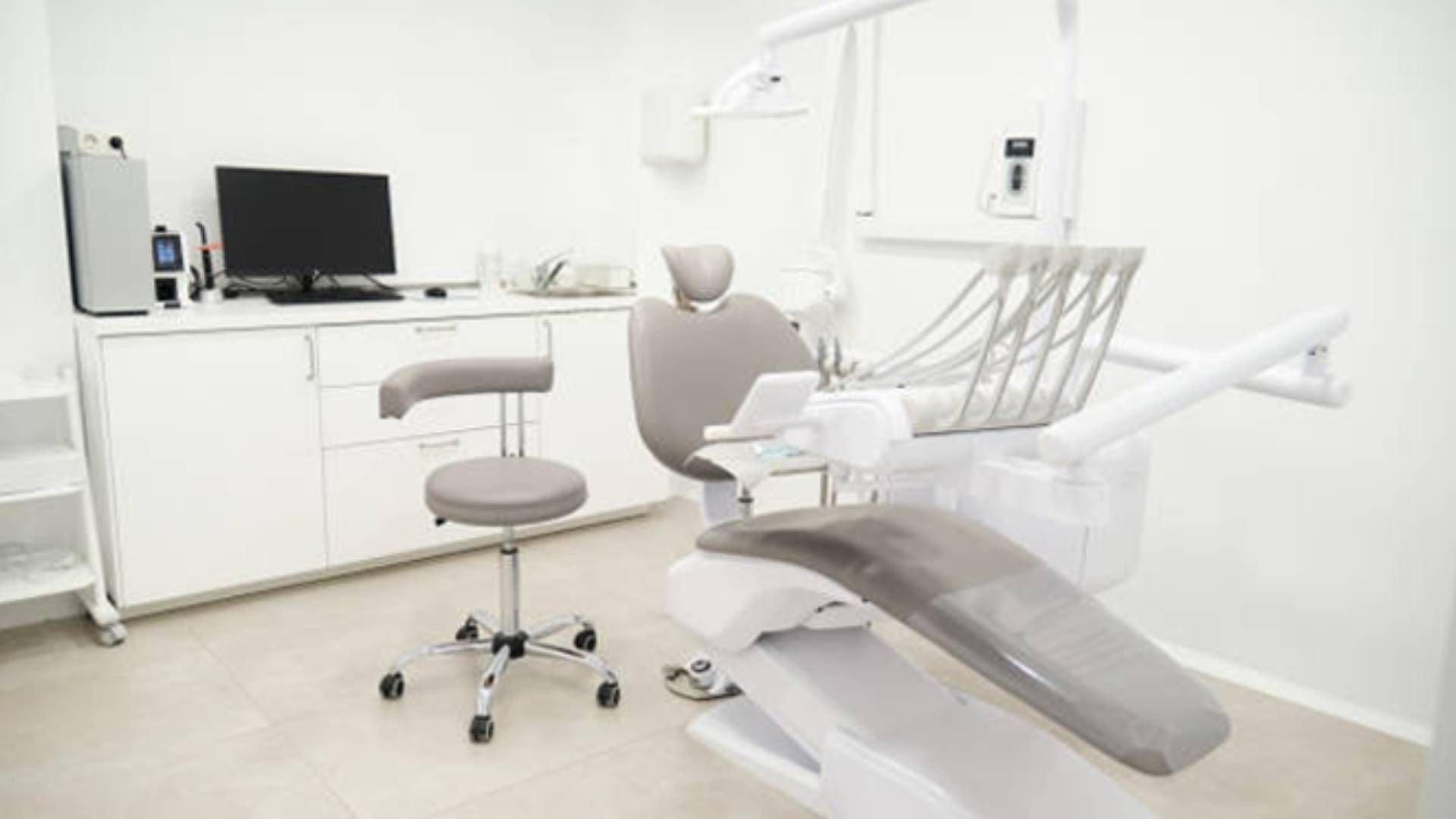 A bright, sterile dental treatment room featuring a beige patient chair, a rolling stool, and a desk with a monitor.