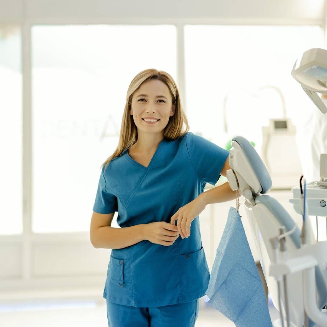 A healthcare professional in blue scrubs smiling while leaning against a dental chair in a bright, modern clinic.