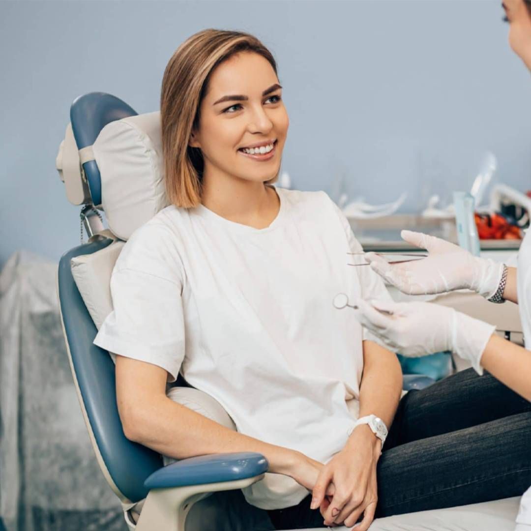 A smiling patient sits in a dental chair while a practitioner in white gloves holds dental tools in a bright office.