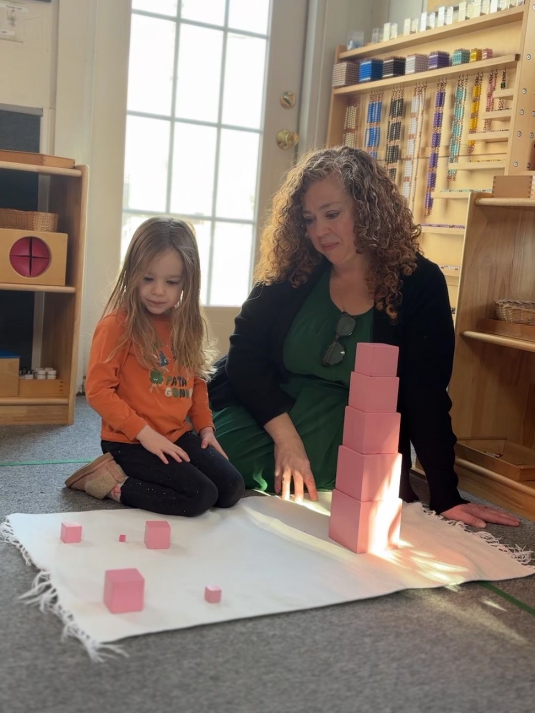 A primary Montessori child and teacher work together to build the pink tower on a floor rug.
