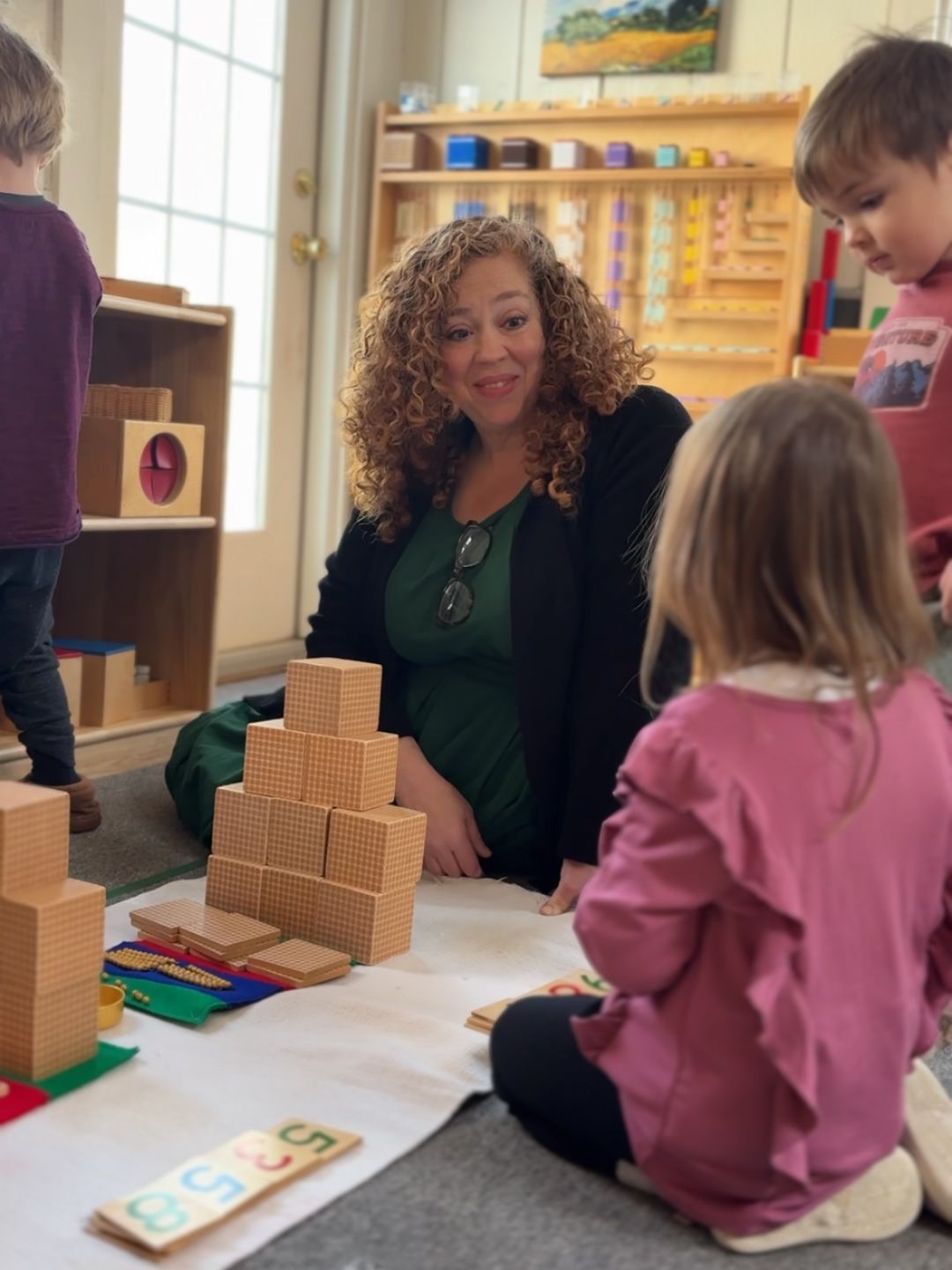 A primary Montessori teacher guides a child through a math lesson using golden bead thousand cubes on a floor rug.