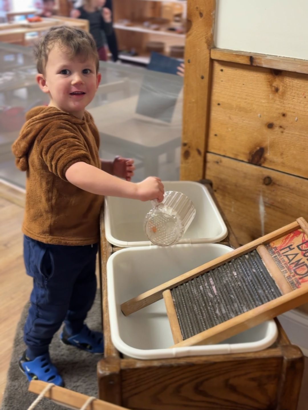 A toddler smiles while pouring water from a pitcher to wash cloth with a washboard in a Montessori classroom.