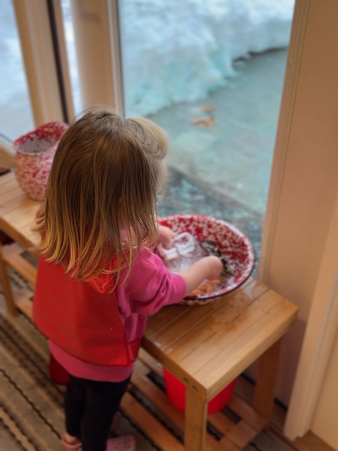 A toddler Montessori child wears an apron while practicing dish washing in a basin at a wooden stand.