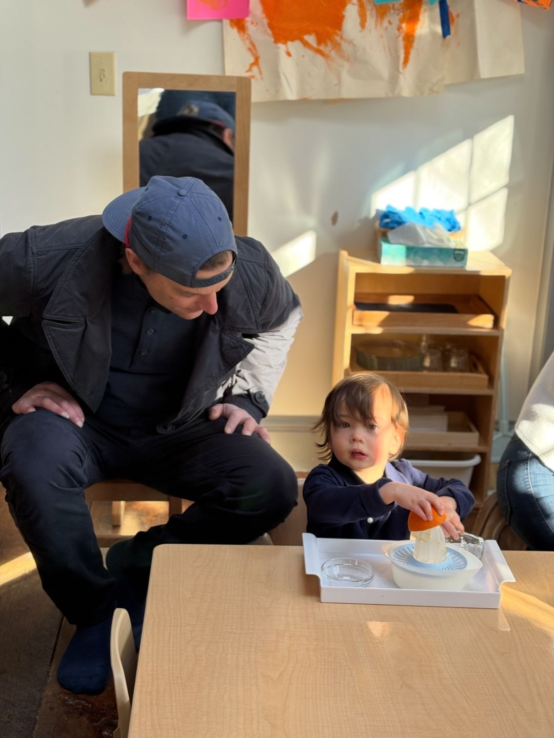 A toddler Montessori child uses a juicer to squeeze an orange while an adult observes in a classroom setting.