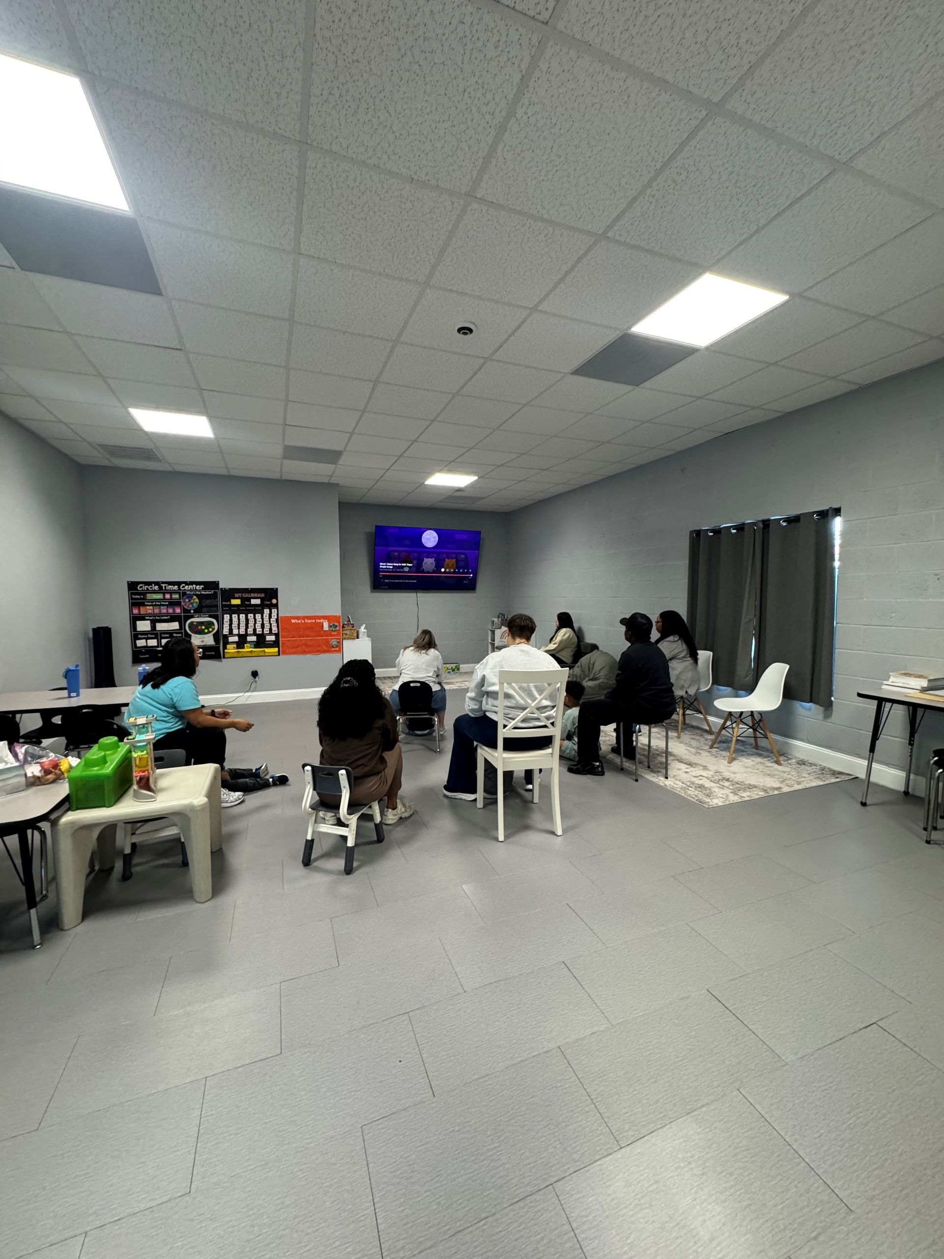 People seated in a room, watching a screen. Grey walls, fluorescent lighting, and some tables.