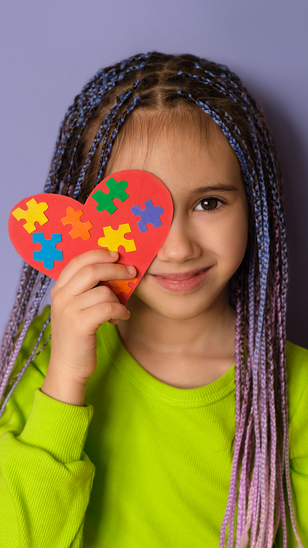 Girl with braided hair holds a heart with puzzle pieces over one eye, smiling; purple background.