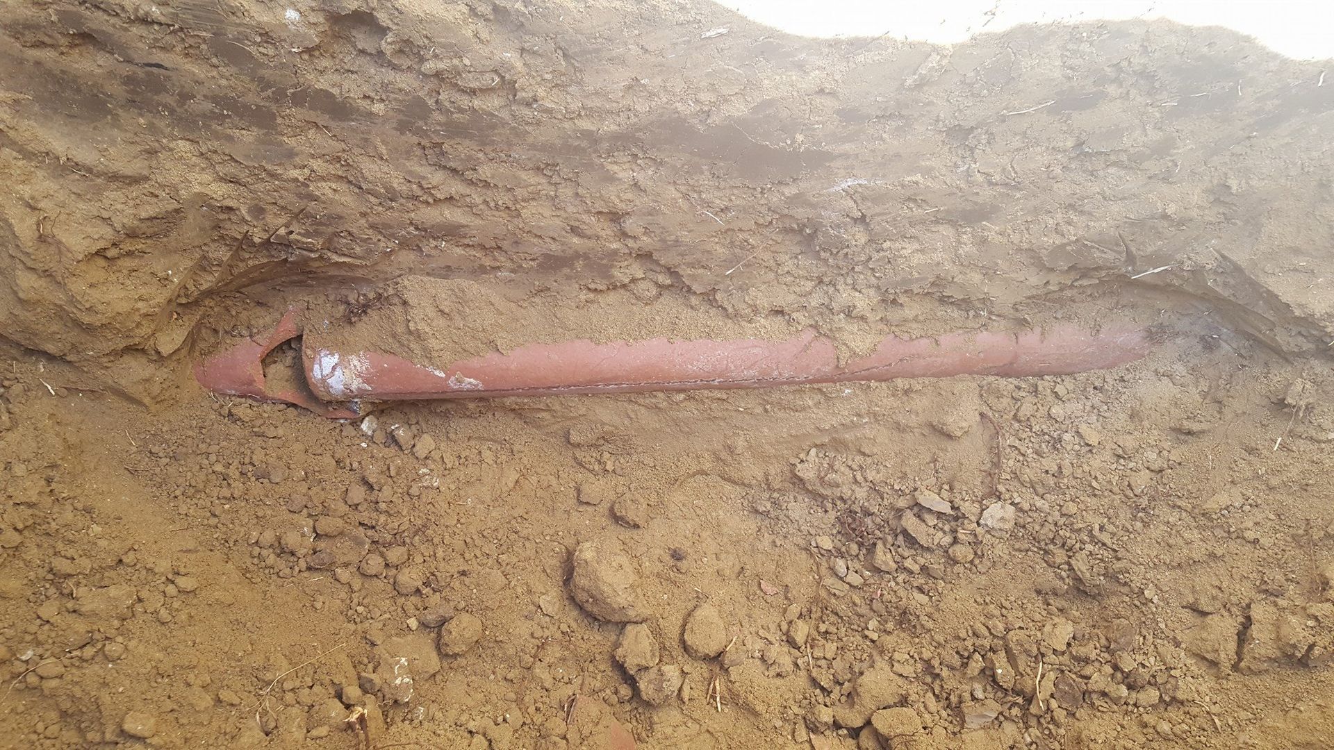 A red pipe is laying in the dirt in a hole.
