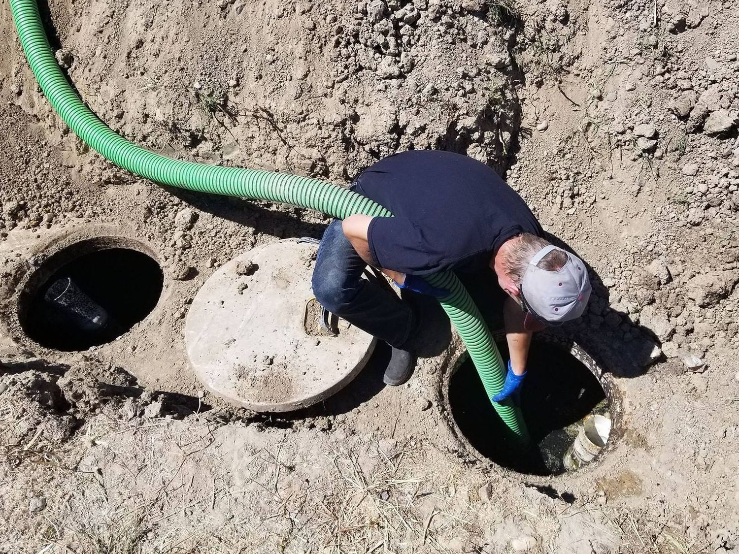 A man is using a green hose to pump water into a septic tank.