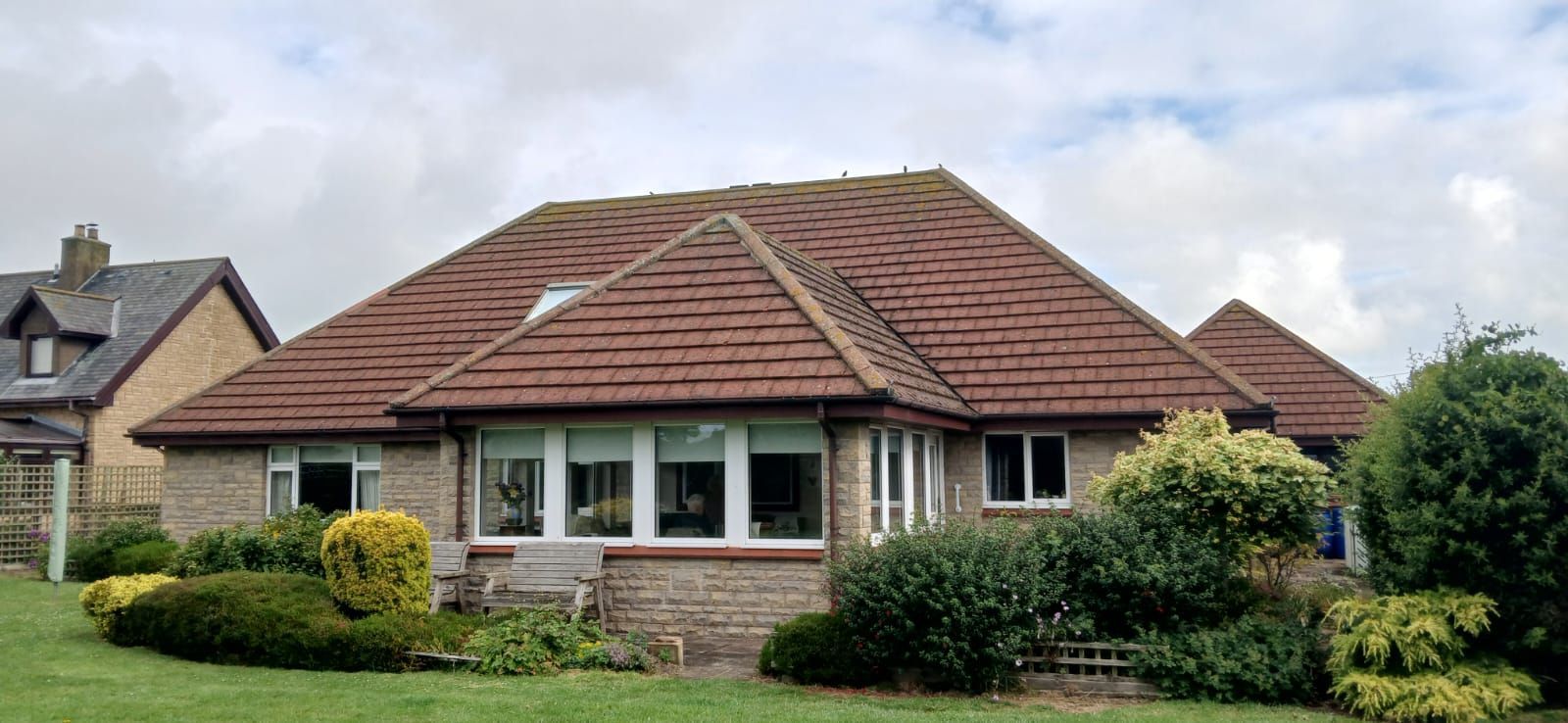 Low brick house with red tiled roof, windows, and greenery in front, cloudy sky.