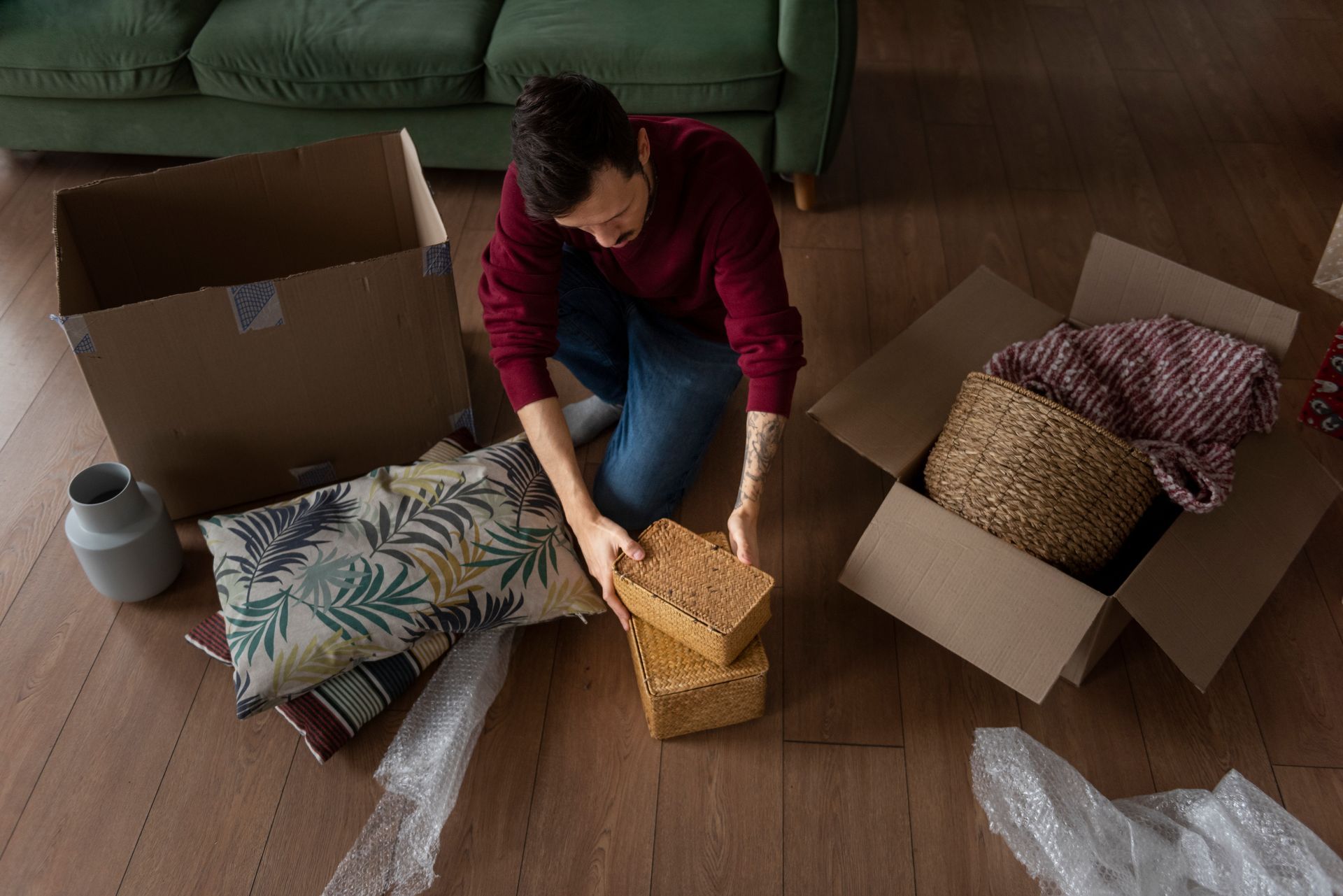 Man kneeling, packing boxes on wooden floor, surrounded by items and a green couch.