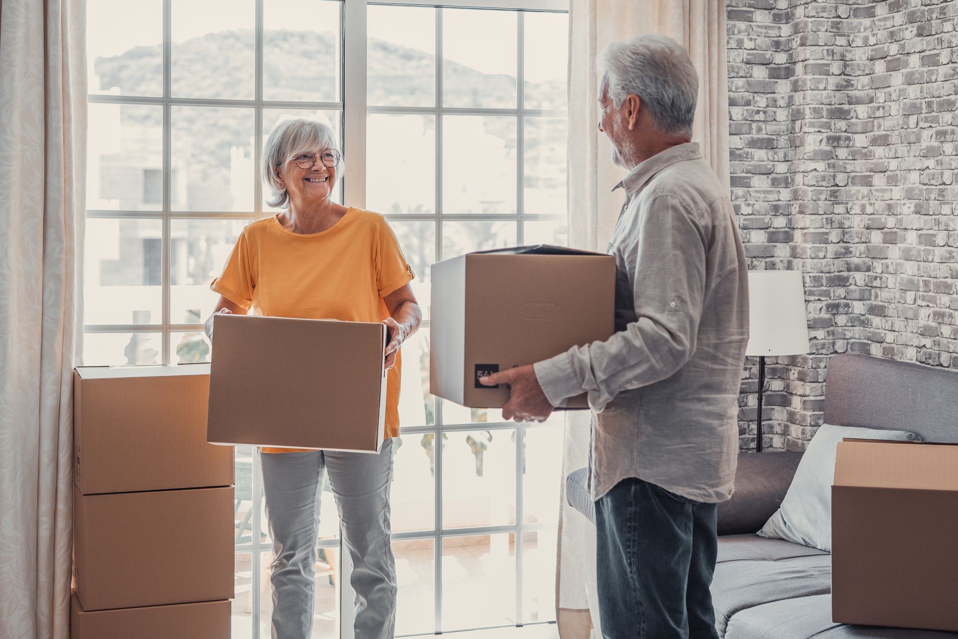 Elderly couple carrying boxes in a room with a window, smiling at each other.