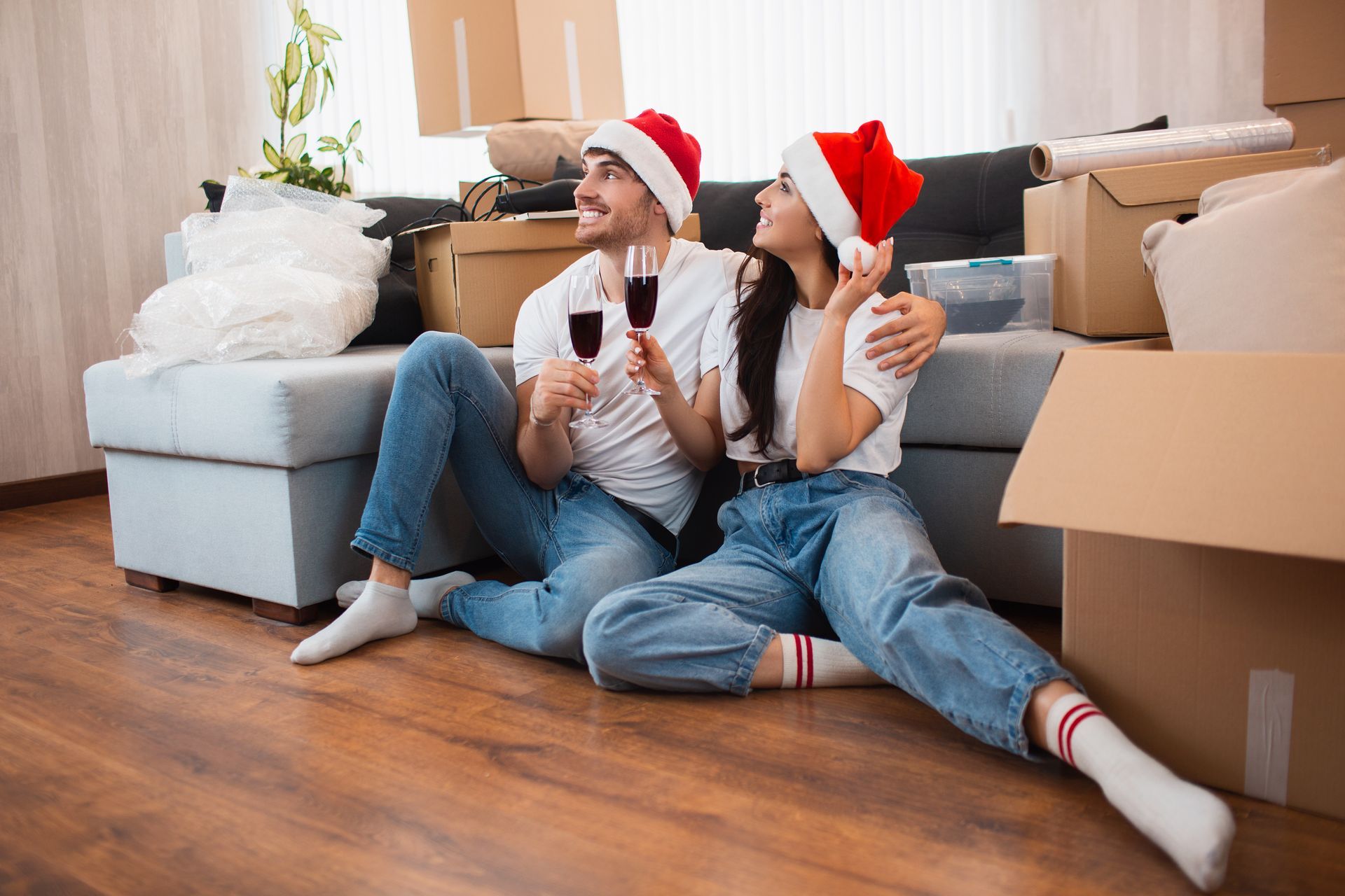 Couple in Santa hats toasting wine, sitting on the floor of a new home among moving boxes.