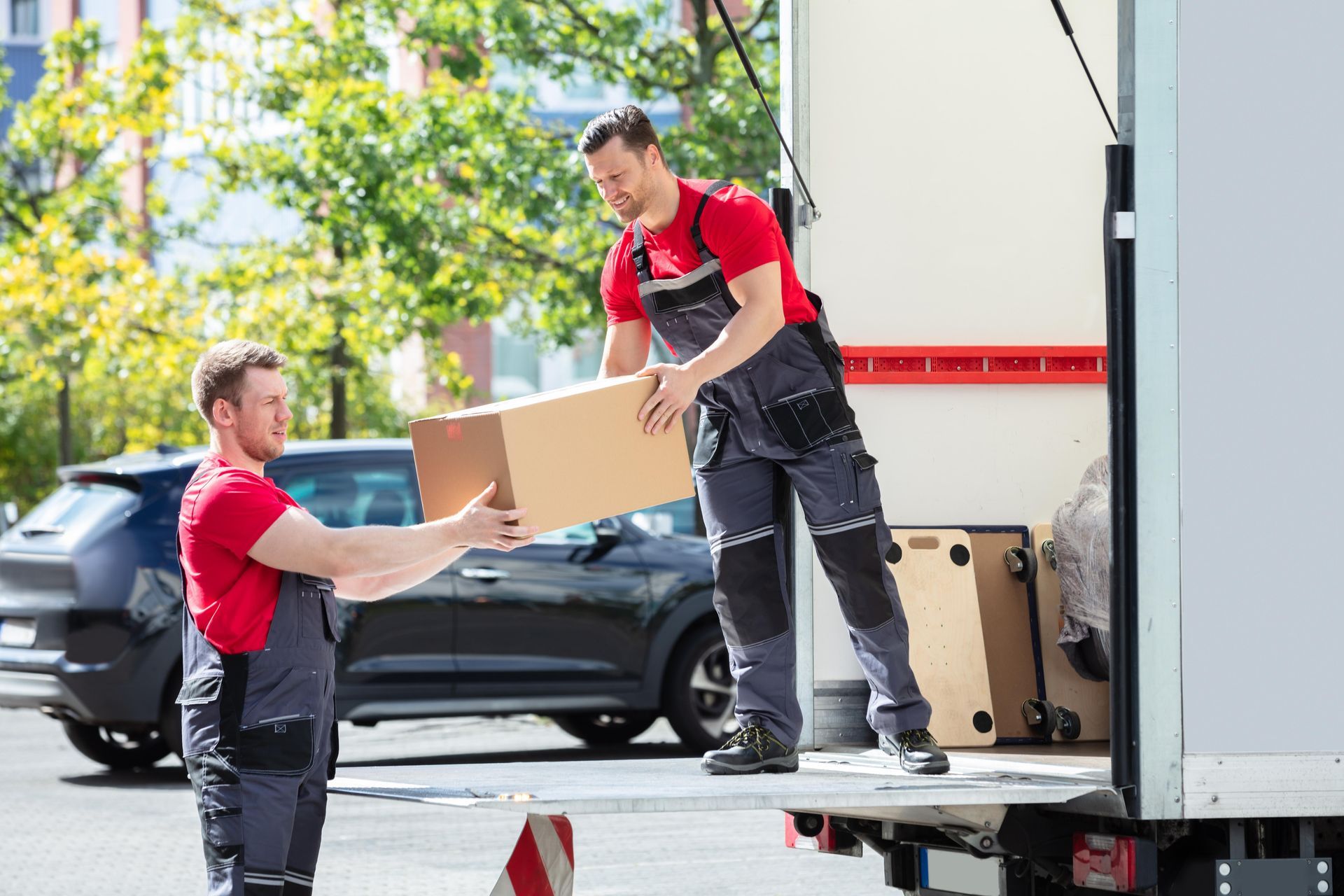 Two movers in red shirts and gray overalls load a cardboard box into a moving truck on a sunny day.