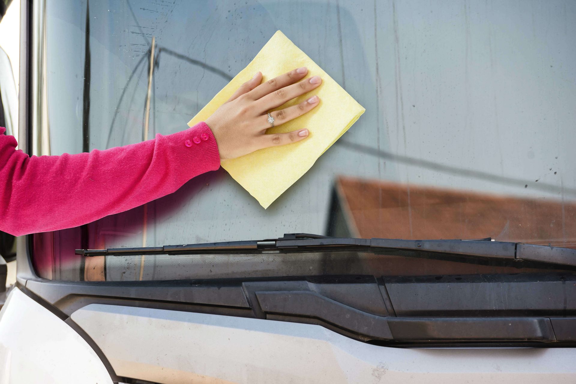 Hand wiping a yellow cloth on a vehicle's windshield. Pink sleeve visible.