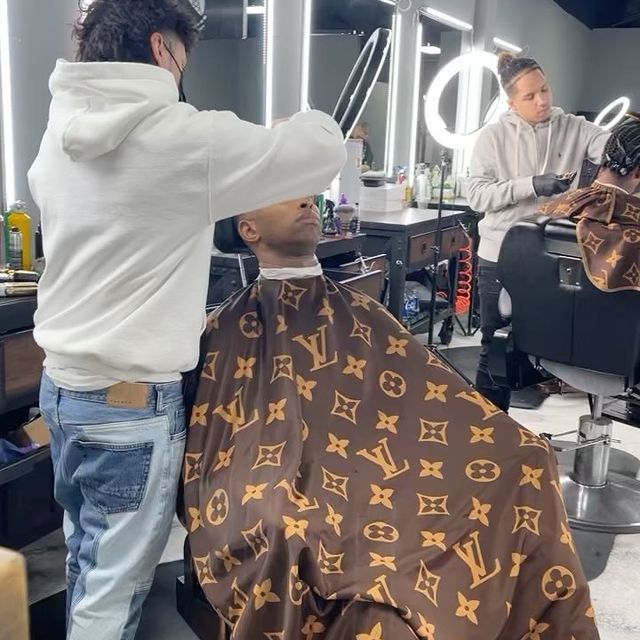 A man is getting his hair cut by a man wearing a louis vuitton cape