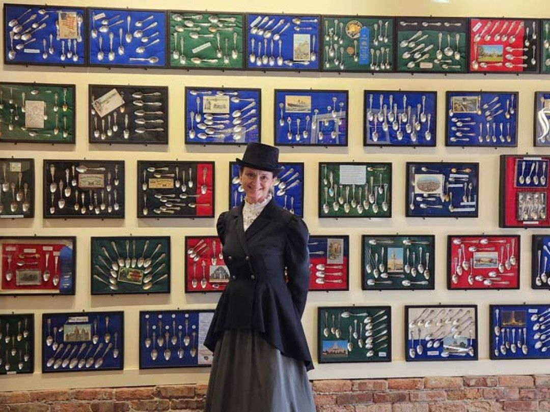 Woman in vintage attire poses before a wall of framed spoons in various colors.