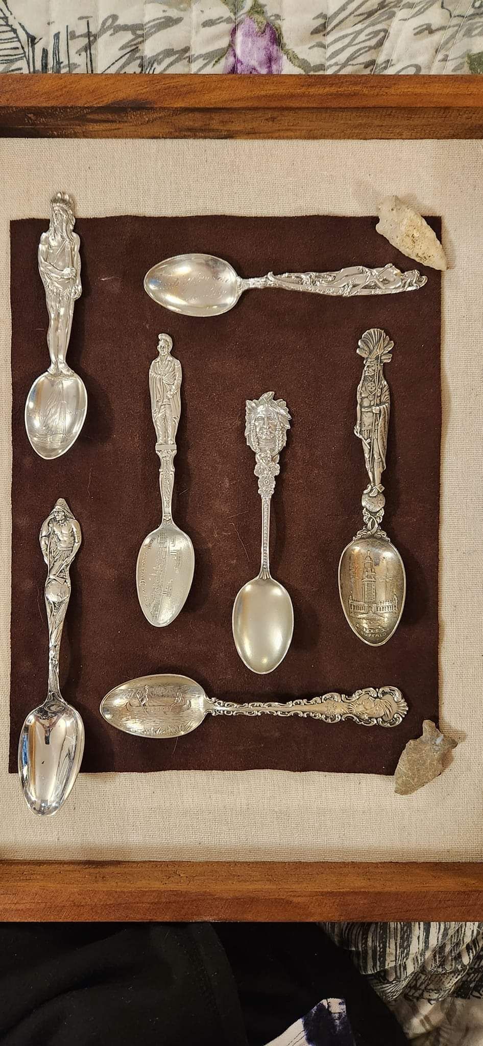Framed display of seven ornate silver spoons on a brown background.