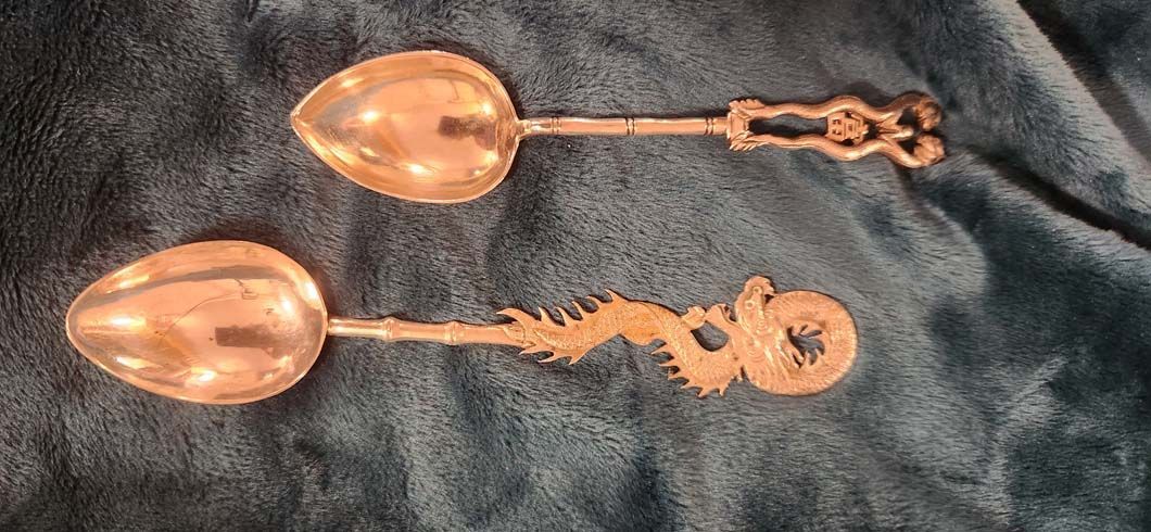 Two silver spoons with decorative handles on a blue surface. One has a dragon handle.