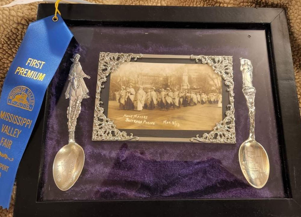 Framed antique display: two silver spoons flank a photo, blue ribbon reads.
