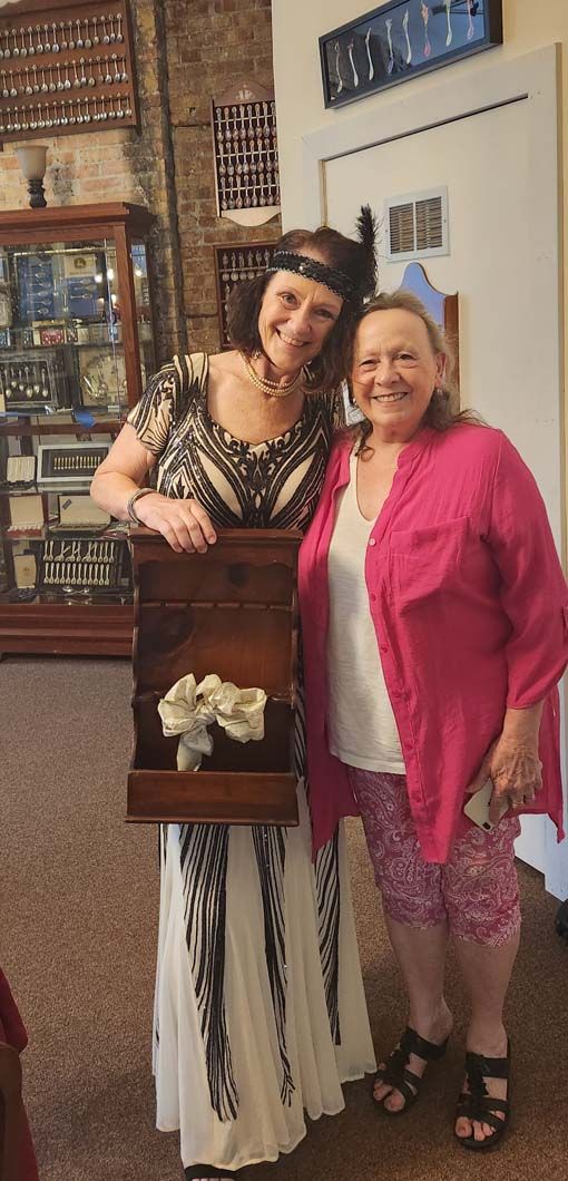 Two women pose; one in costume holding a wooden box with flowers. 