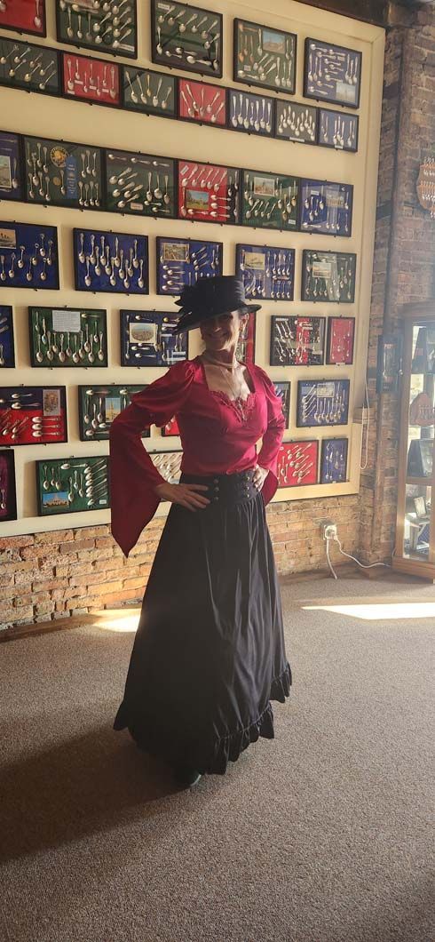 Woman in a red blouse and black skirt, with a hat, poses in front of a wall of framed items.