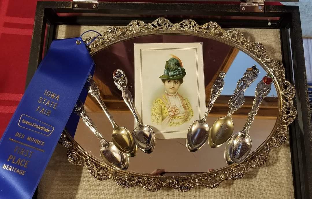 Display case with ornate silver spoons, portrait, and blue ribbon.