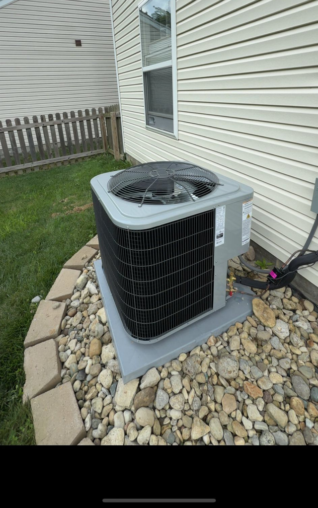 Outdoor air conditioner unit beside a house, with a garden hose nearby and rocks around the base.