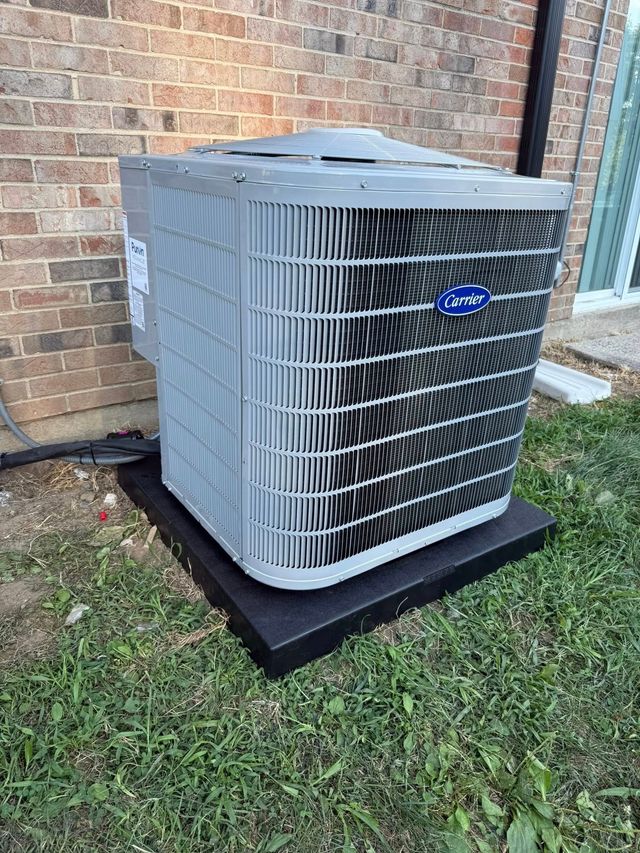 Gray outdoor air conditioner unit on a black pad beside a brick wall and grass.