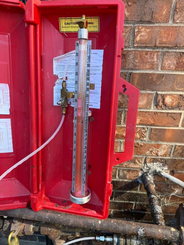 Red fire safety cabinet with a long metal extinguisher mounted inside against a brick wall.
