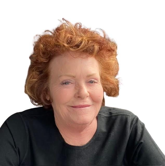 A smiling person with shoulder-length curly red hair wears a black long-sleeved shirt in a restaurant.