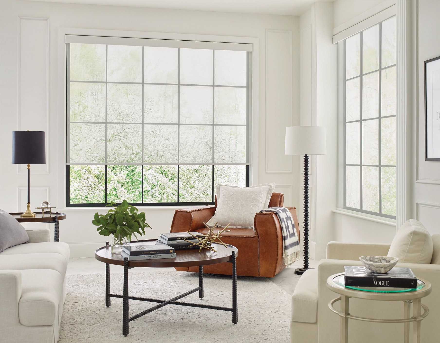 A bright, modern living room with neutral-toned furniture, a round coffee table, and large windows with grid muntins.