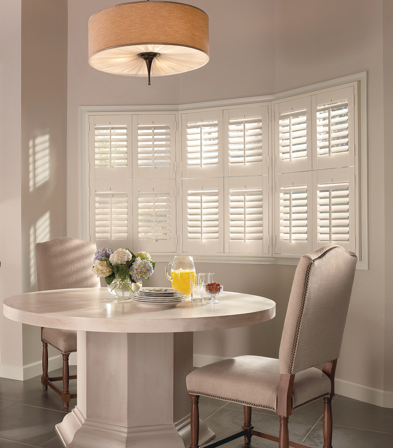 A dining table and two chairs set in front of a bay window with white plantation shutters and a fabric drum pendant light.