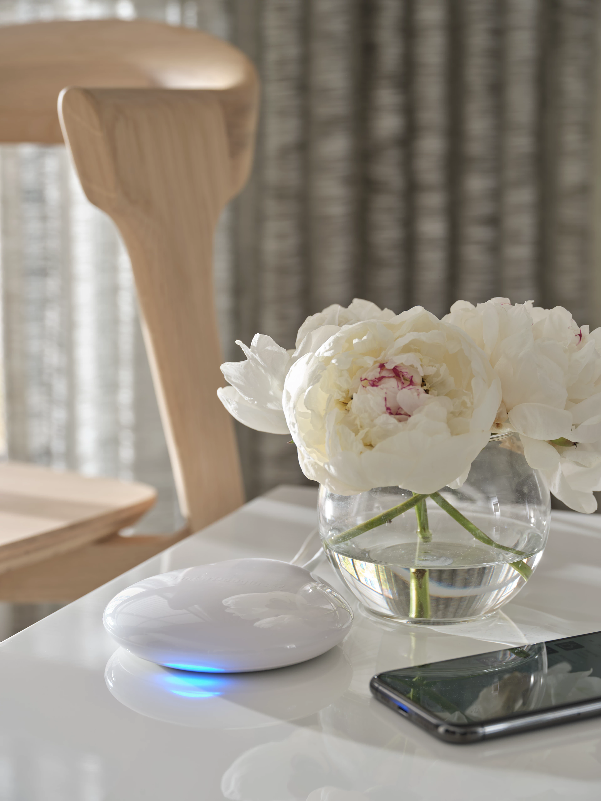 A glowing white tech device, a vase of white peonies, and a smartphone on a white surface next to a wooden chair.